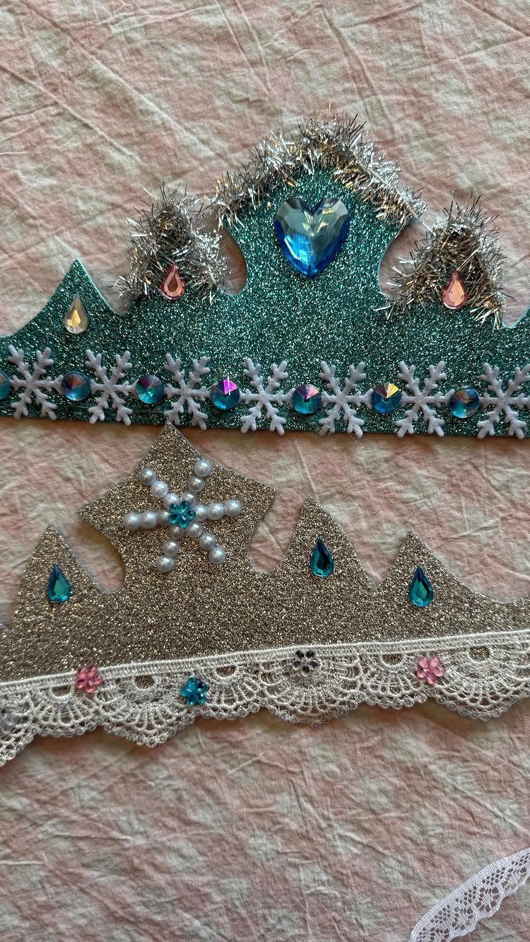 Two glittery Frozen-themed crowns decorated with blue gems and snowflakes on pink fabric.