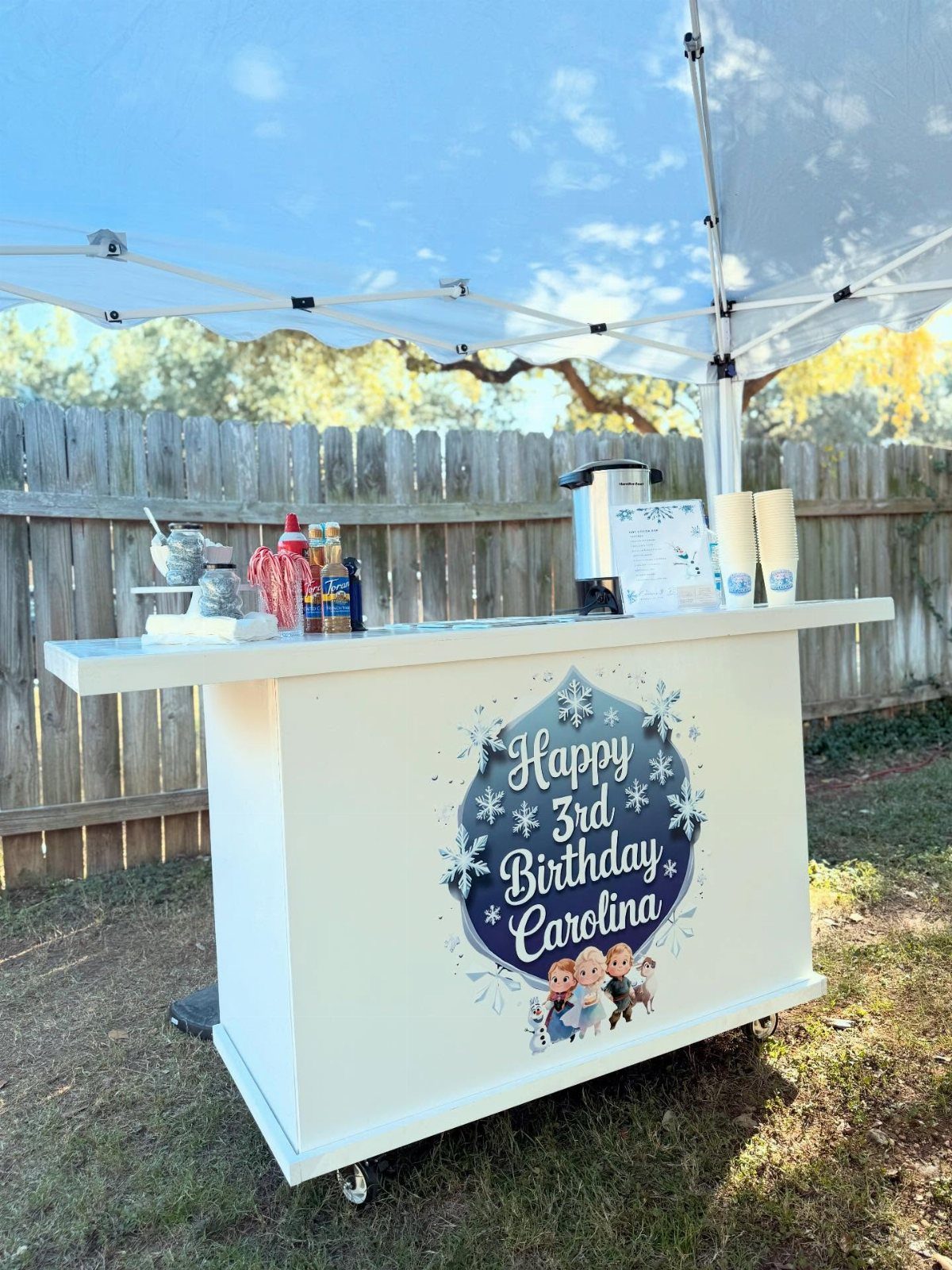 White beverage station with a Frozen birthday sign and hot cocoa supplies outdoors.