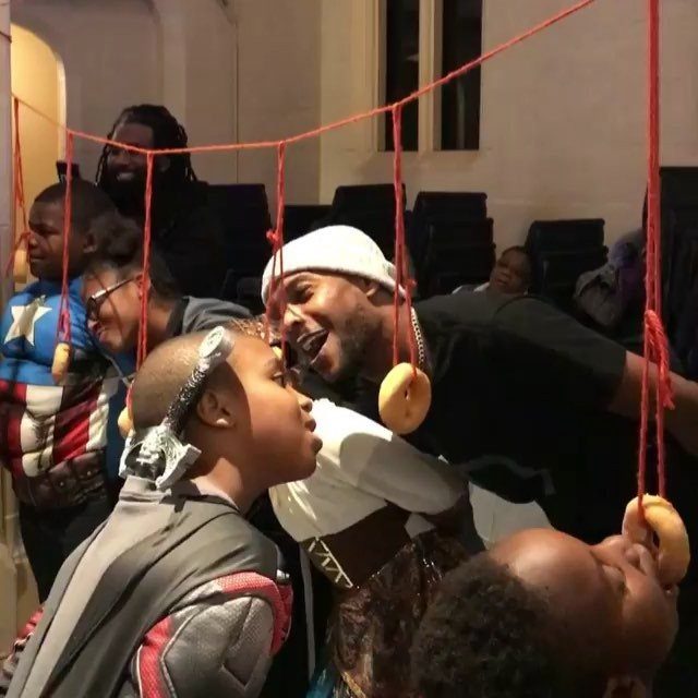 People in costumes trying to eat donuts hanging from red strings indoors.