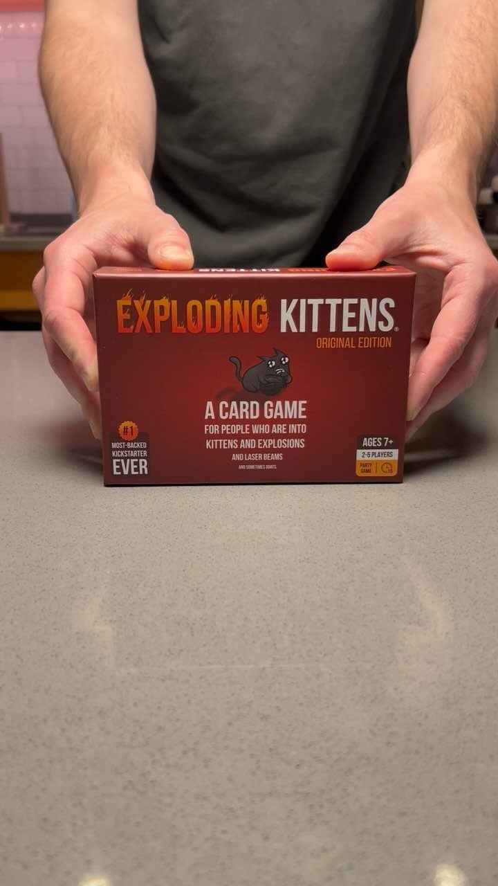 Exploding Kittens card game box held by a person over a grey table