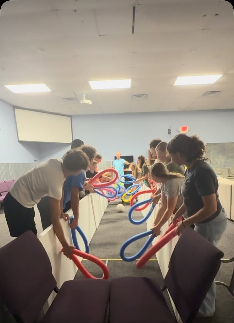 Teenagers using colorful pool noodle loops to transport a ball across a room.