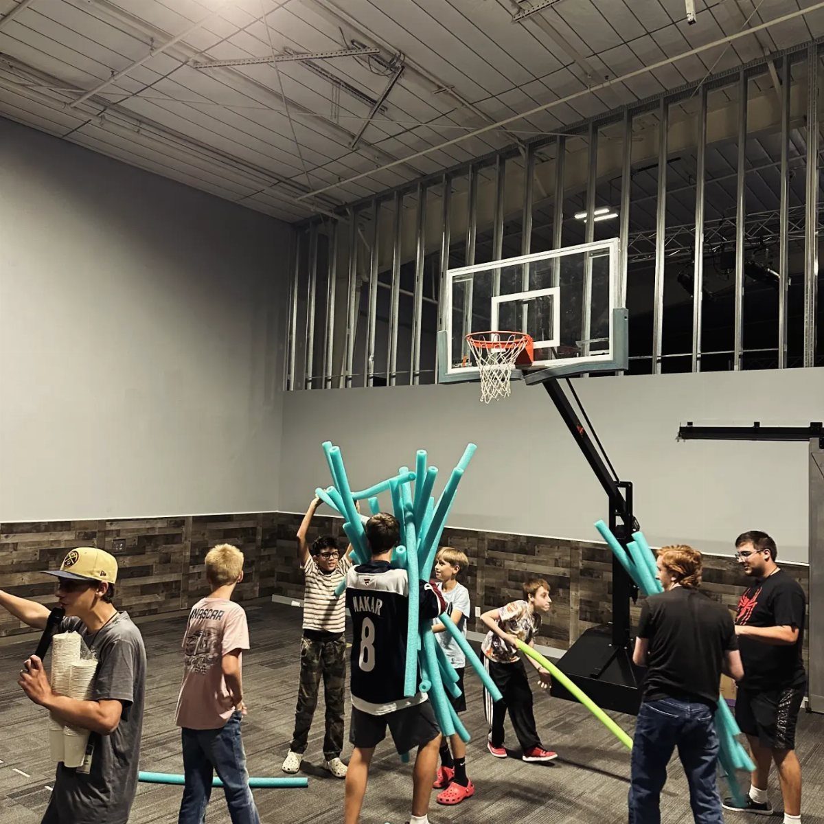 Teenage boys playing with blue and yellow pool noodles in a gym.