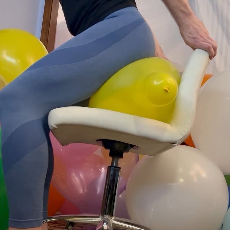 Person sitting on a white chair popping a yellow balloon with their body