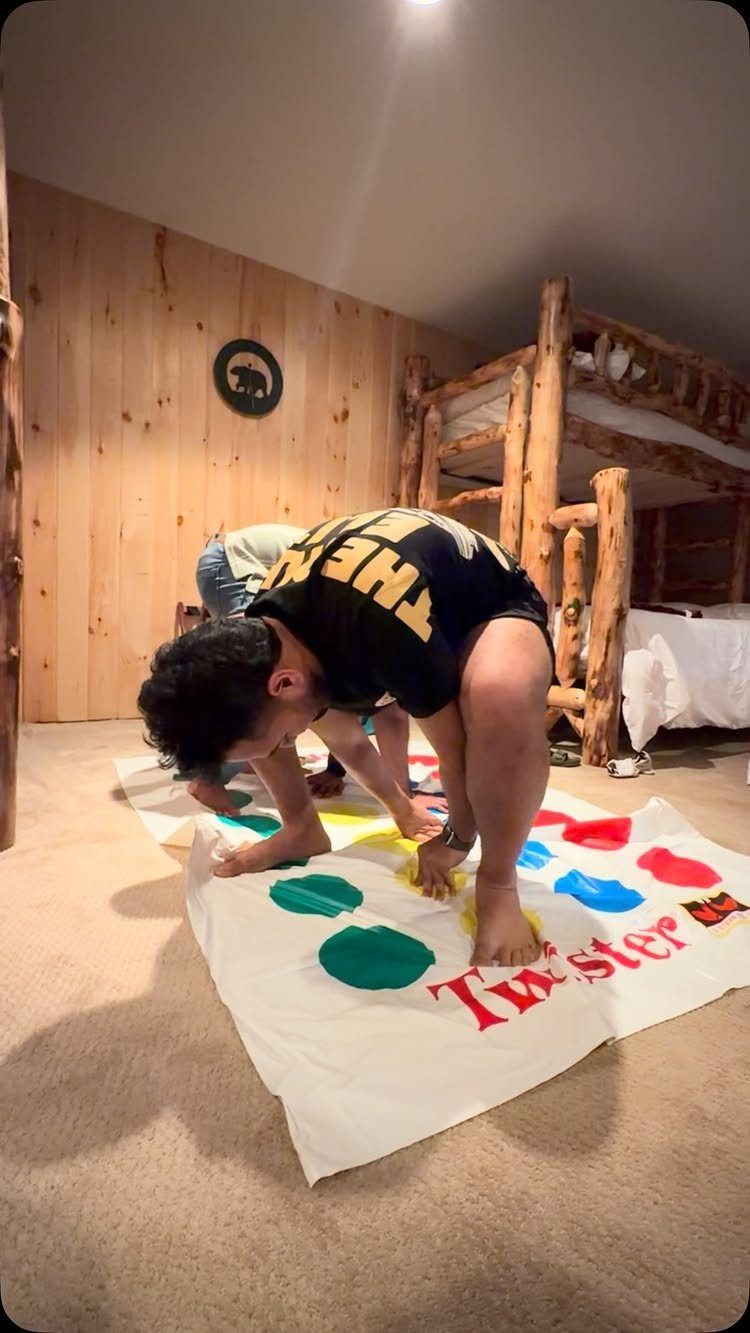 Two people bending over while playing Twister on a mat in a rustic room.
