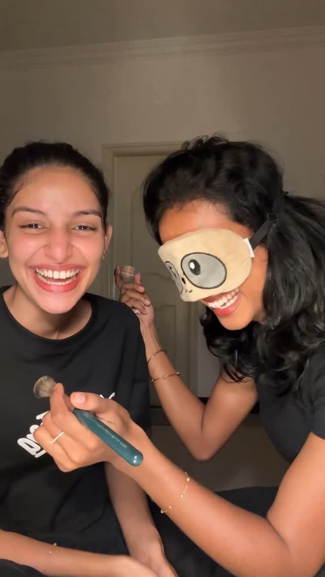 Woman wearing a sleep mask applying makeup to a laughing friend with a brush.