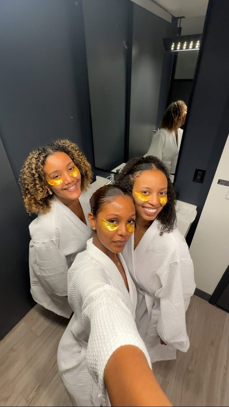 Three women in white robes wearing yellow under-eye masks posing for a selfie.