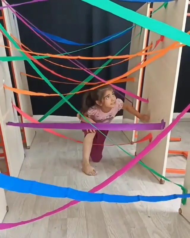 Young girl crawling through a colorful ribbon obstacle course between white walls indoors.