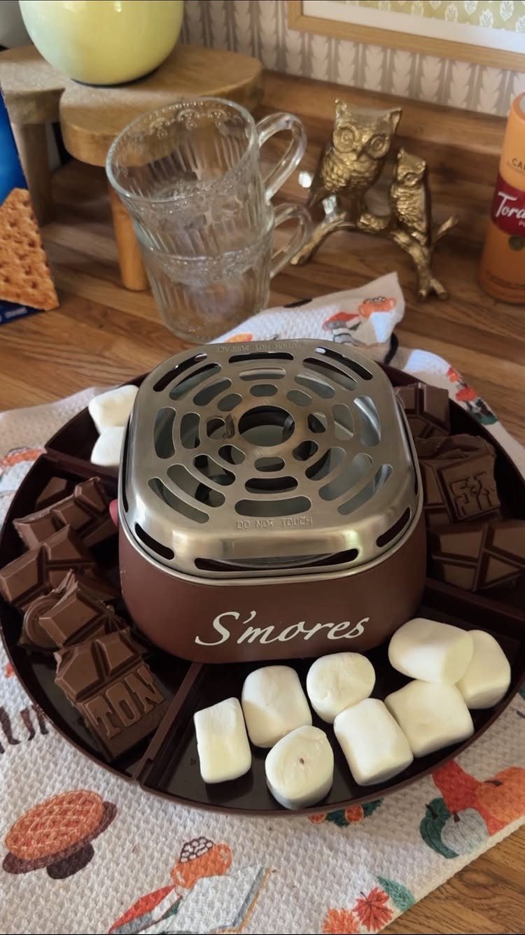 Indoor s'mores maker with chocolate and marshmallows on a wooden table.