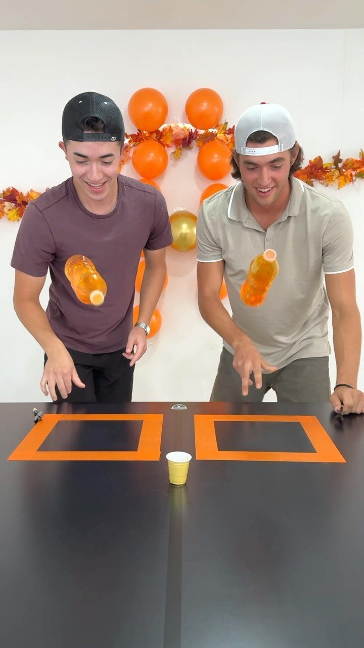 Two young men tossing orange bottles toward targets on a black table.