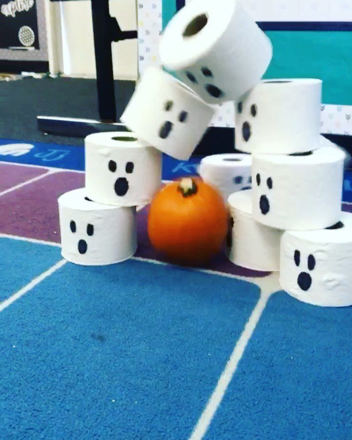 Orange pumpkin knocking over toilet paper rolls decorated as ghosts for bowling game.
