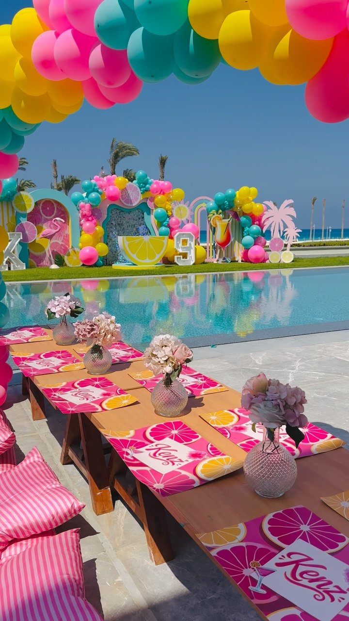 Poolside birthday party setup with colorful balloon arch and themed table decorations.