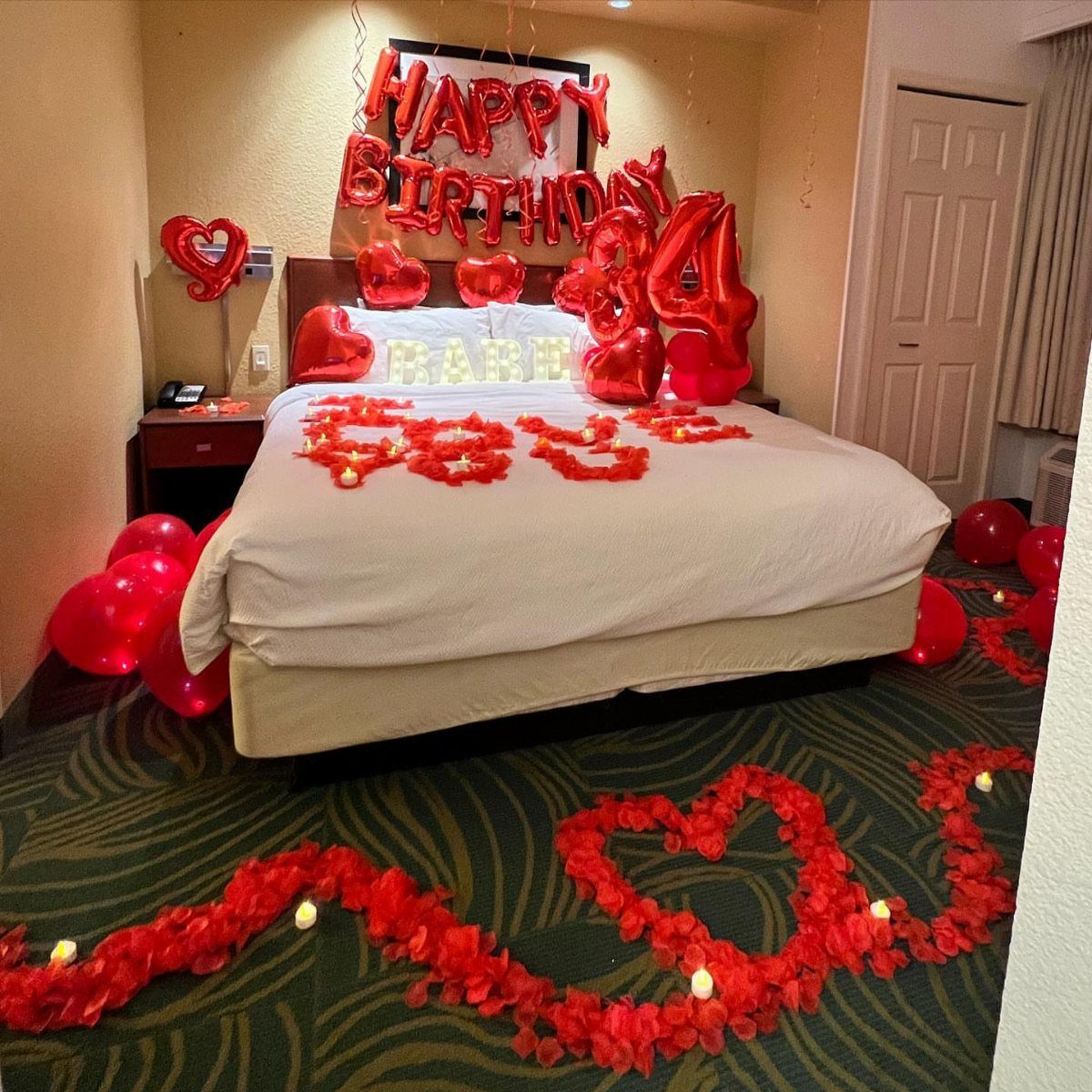 Hotel bed decorated with red rose petals and red birthday balloons.