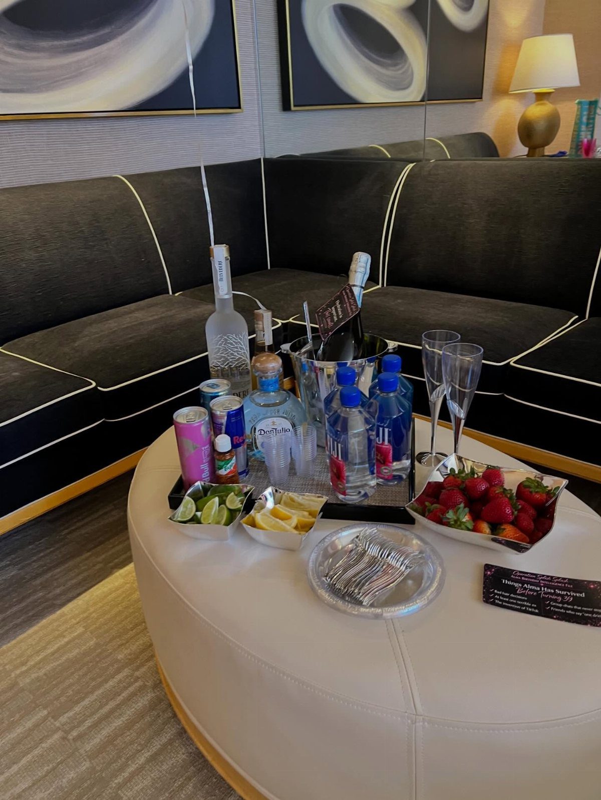 Party spread with champagne, cocktails, and strawberries on a white hotel ottoman table.