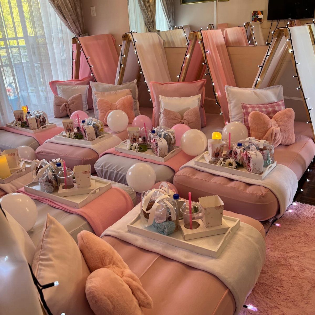 Pink and white teepee sleepover setup with mattresses, balloons, and gift trays.