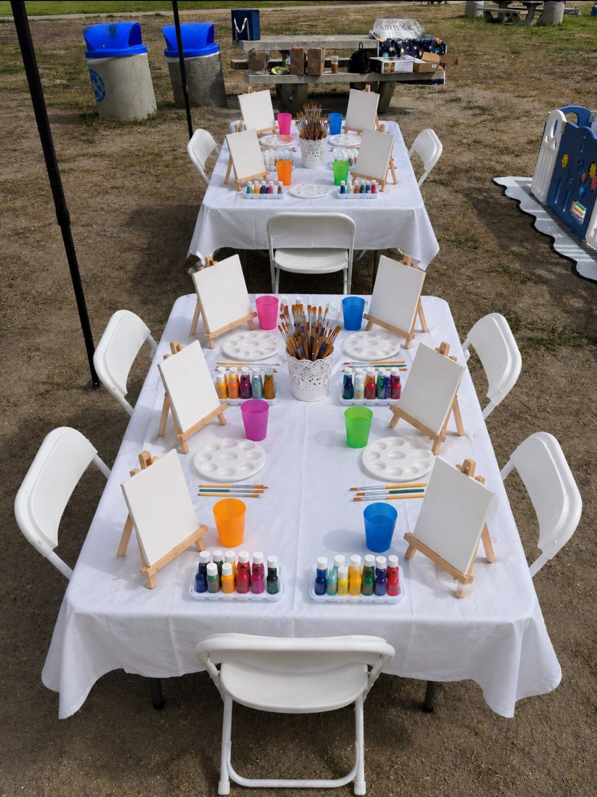 Outdoor art party setup with canvases on easels and paints on white tables.