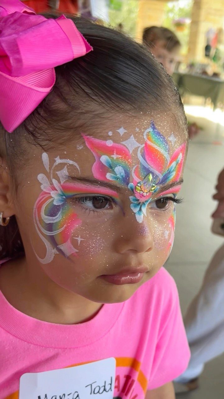Young girl with a pink bow and colorful glittery butterfly face painting.