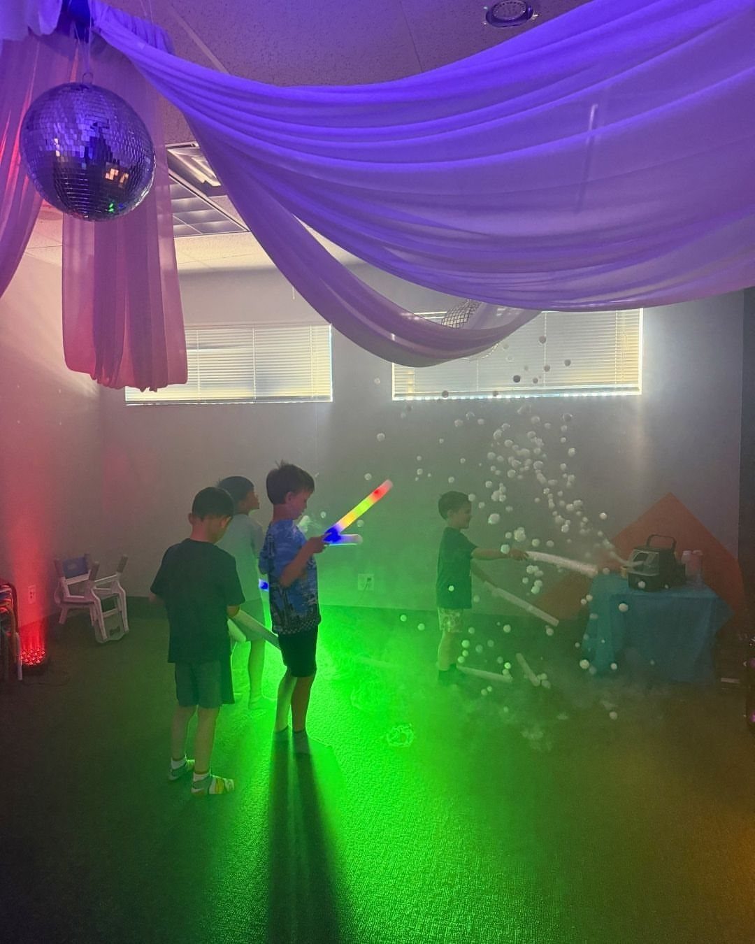 Children playing with light sticks under a disco ball and falling white balloons.