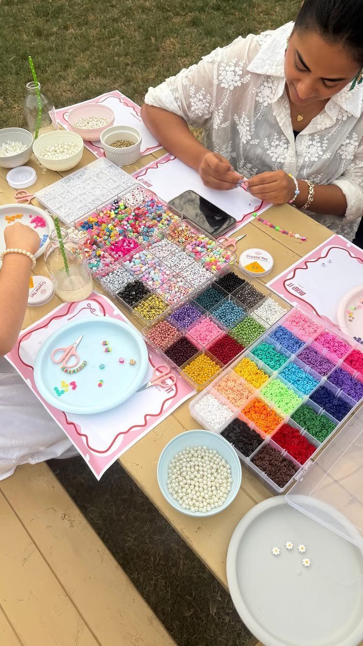 Woman and child making jewelry with colorful beads at an outdoor table.