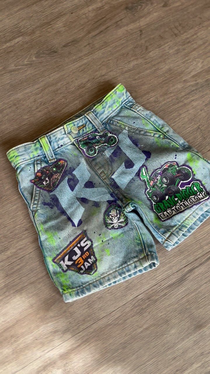 Customized denim shorts with monster truck patches and colorful paint splatters on wood.