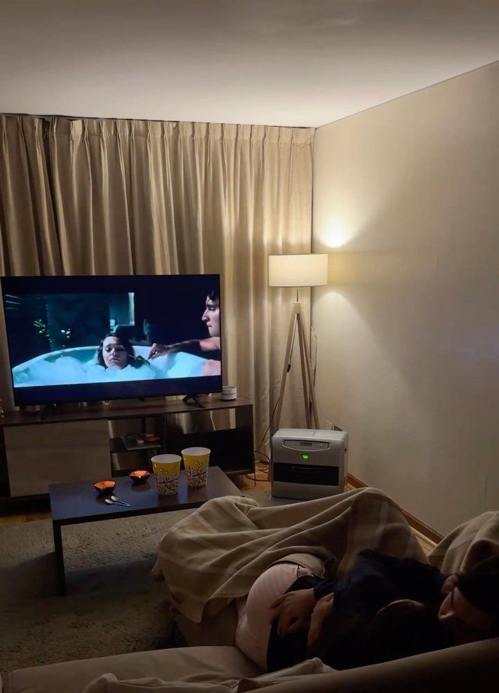 Person under a blanket watching a movie on a TV with popcorn nearby.