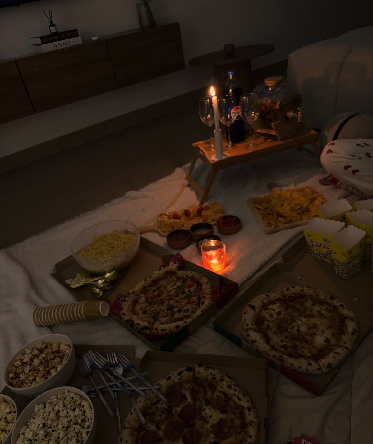 Pizza, popcorn, and drinks spread across a white blanket in a cozy room.