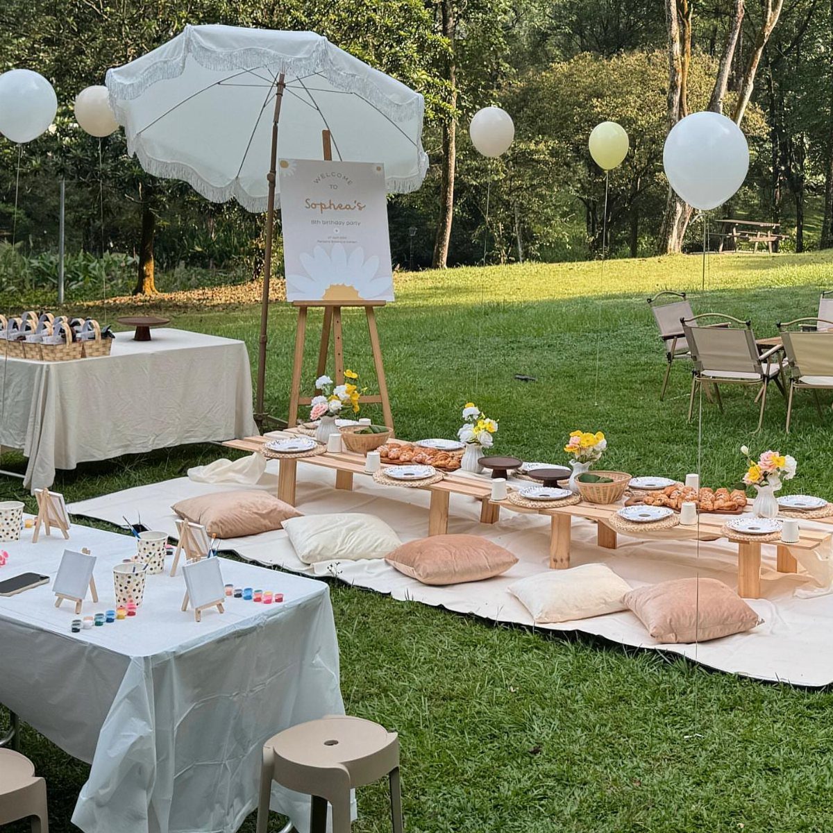 Outdoor party setup with low picnic table, white balloons, and white umbrella on grass