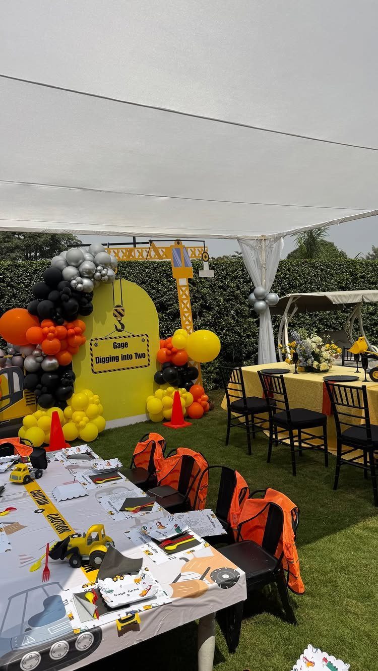 Construction themed outdoor party setup with yellow balloons, toy trucks, and safety vests.