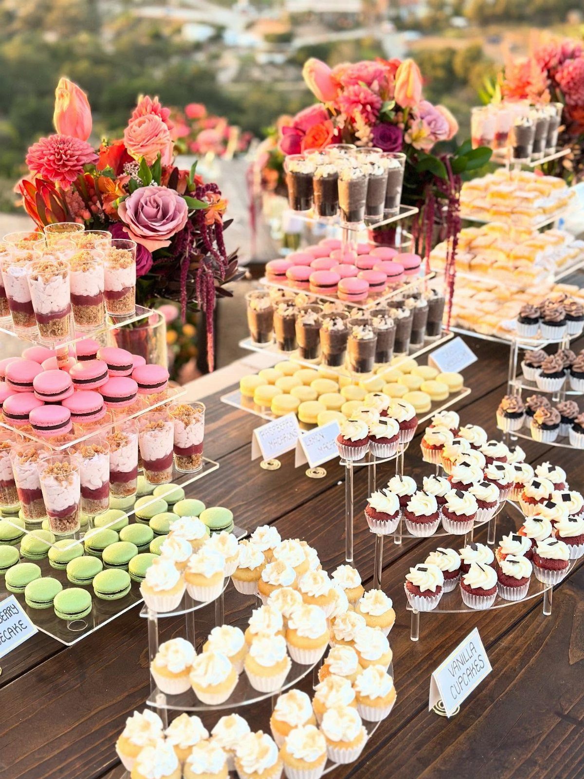Dessert table with macarons, cupcakes, and mousse cups decorated with colorful flowers outdoors.