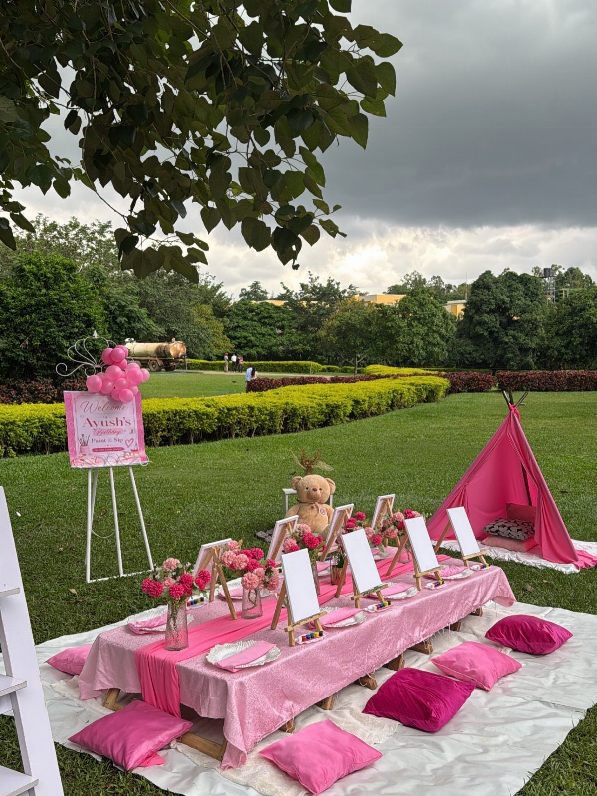 Pink themed outdoor party setup with a long table, teepee, and cushions on grass.