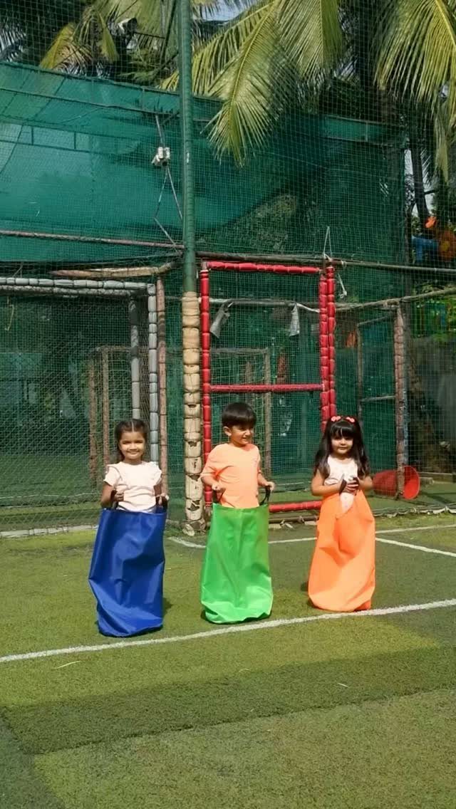 Three children in blue, green, and orange sacks preparing for a sack race outdoors.