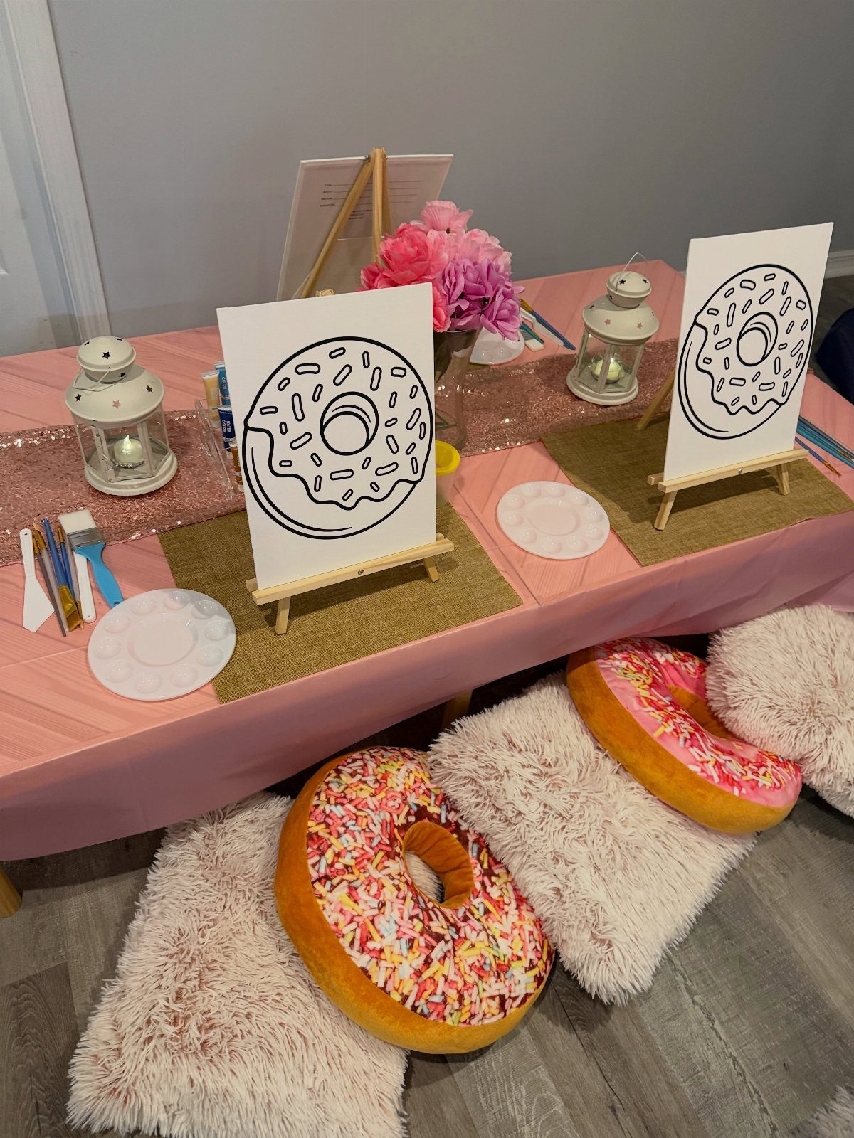 Craft station with donut painting canvases, paintbrushes, and donut pillows on a pink table.