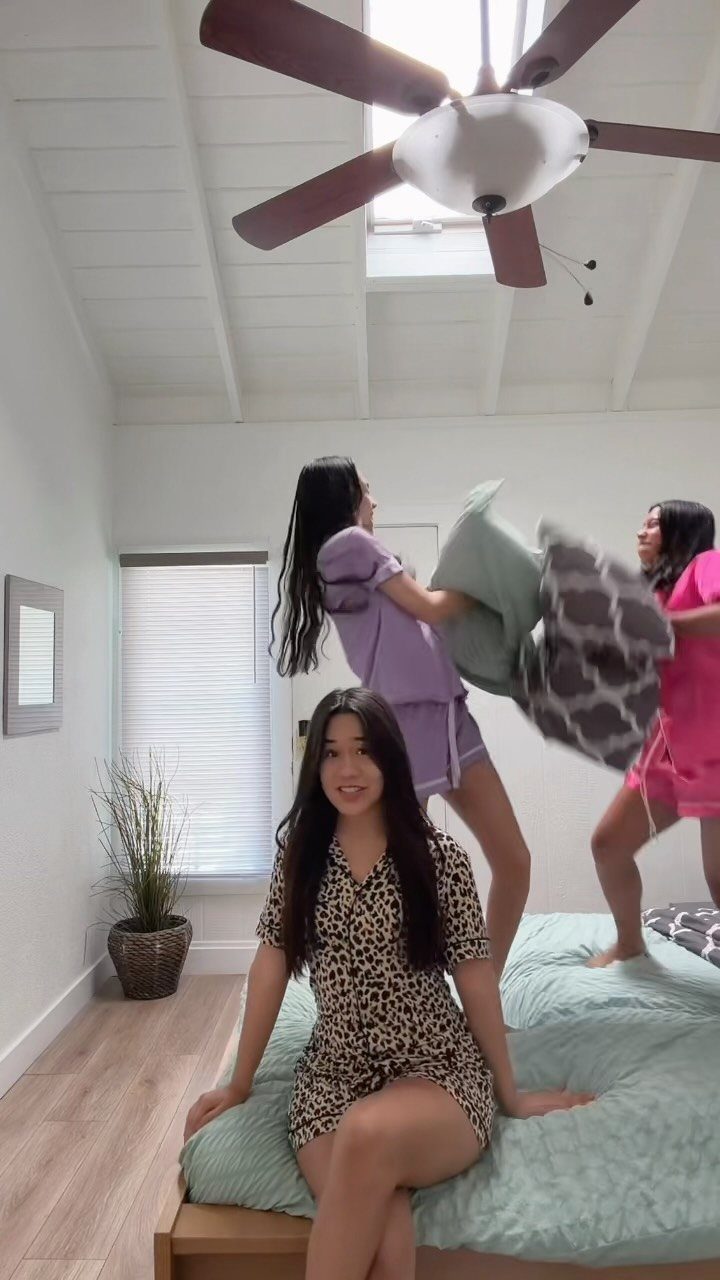 Three women in pajamas, two having a pillow fight on a bed.