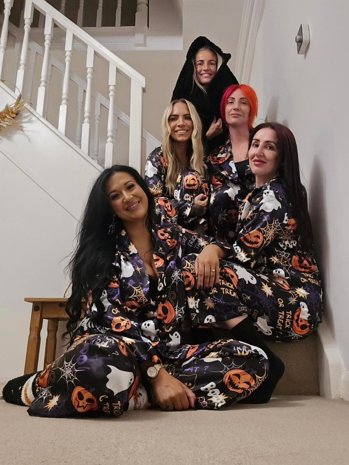 Five women in pumpkin and ghost patterned pajamas posing on a white staircase.