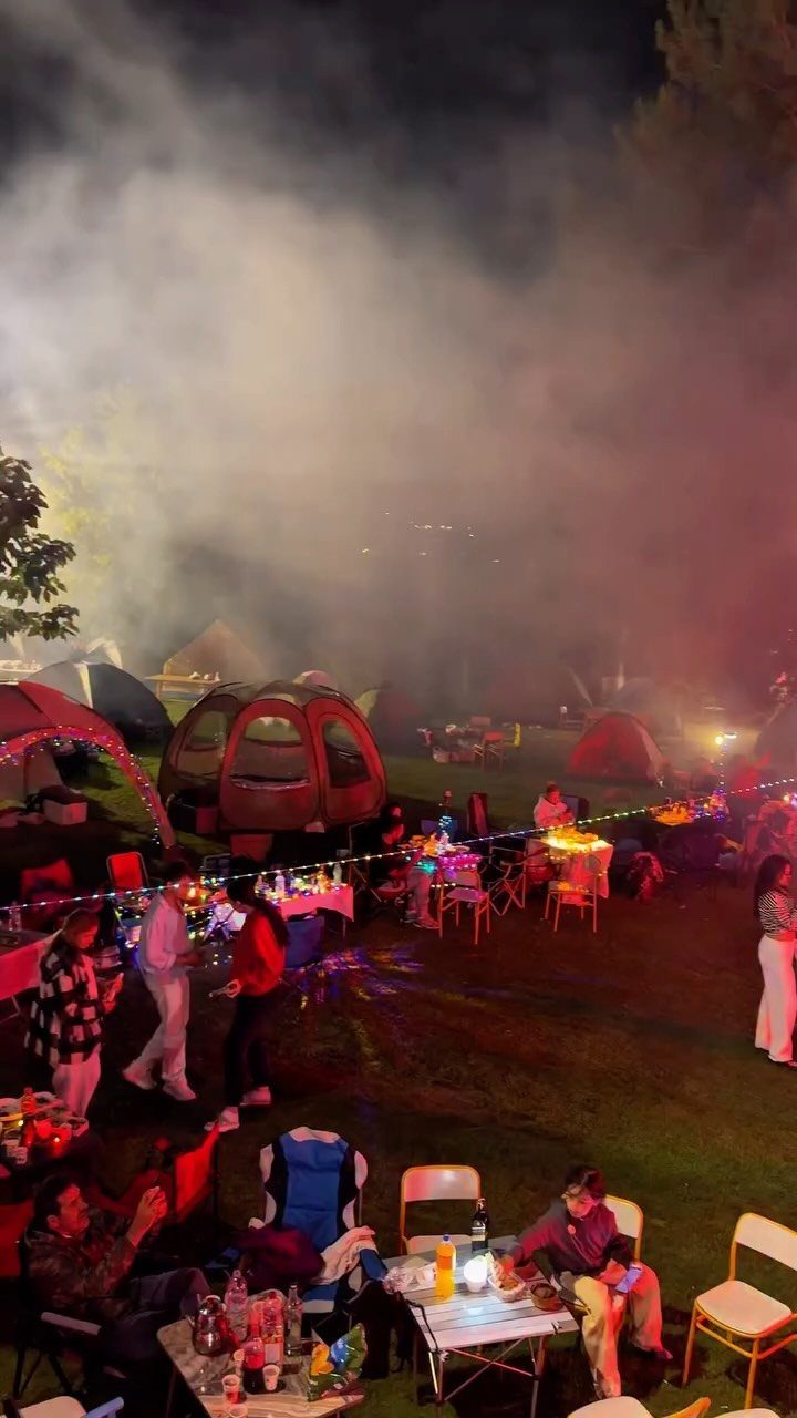 Nighttime camping party with tents, colorful string lights, and people gathering outdoors.