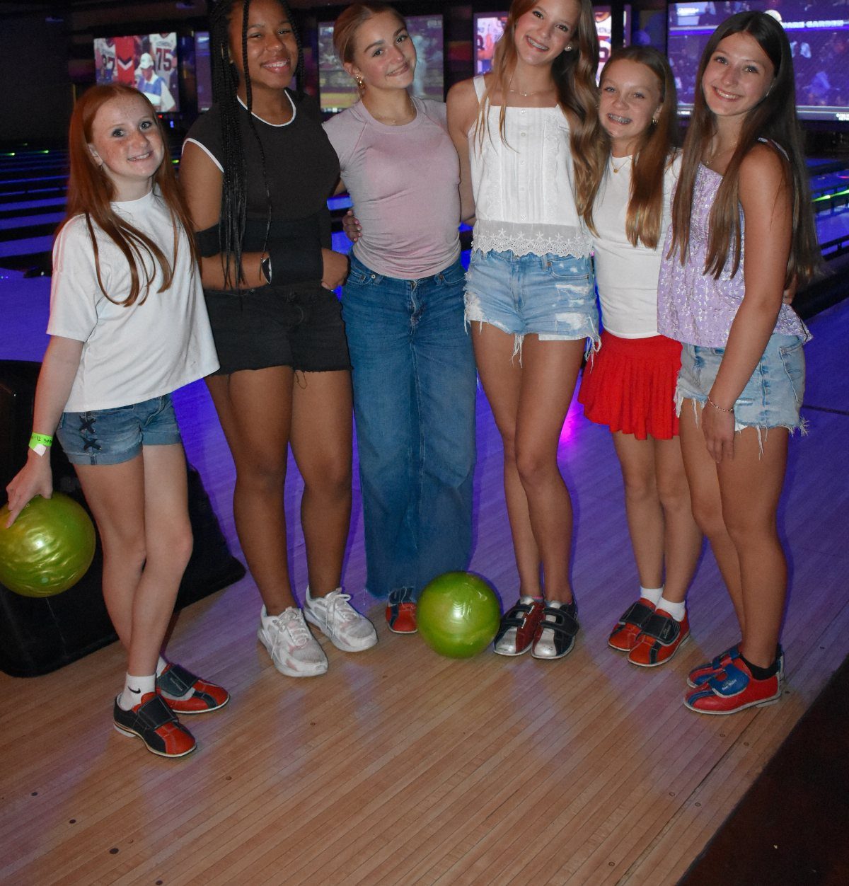 Six teenage girls posing with bowling balls on a wooden bowling alley lane.