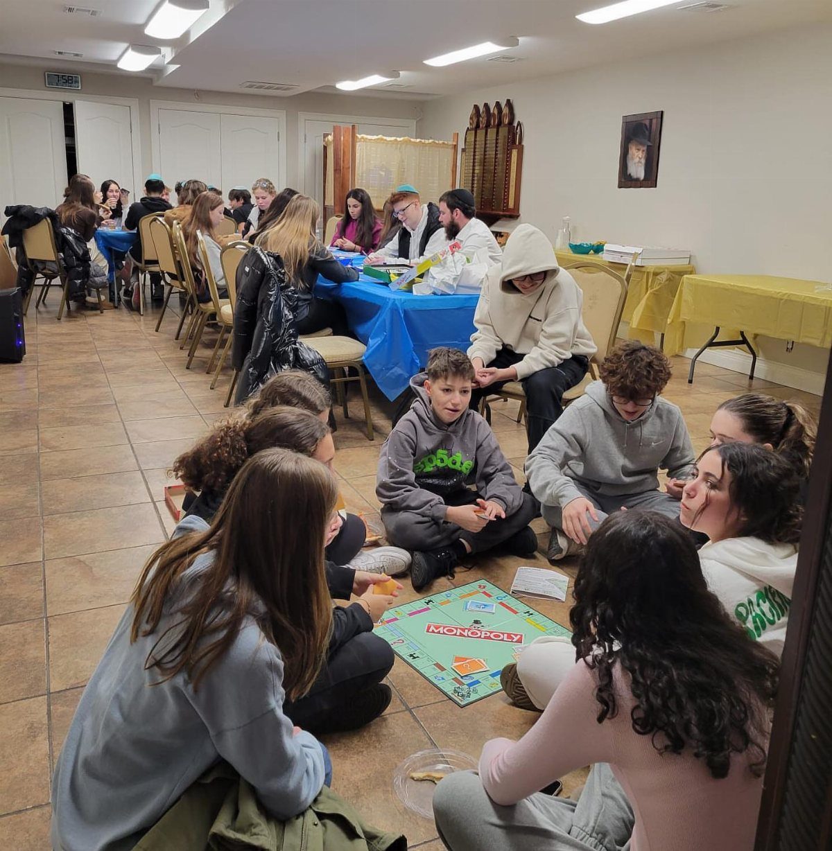 Teenagers sitting on a tiled floor playing a Monopoly board game together indoors.