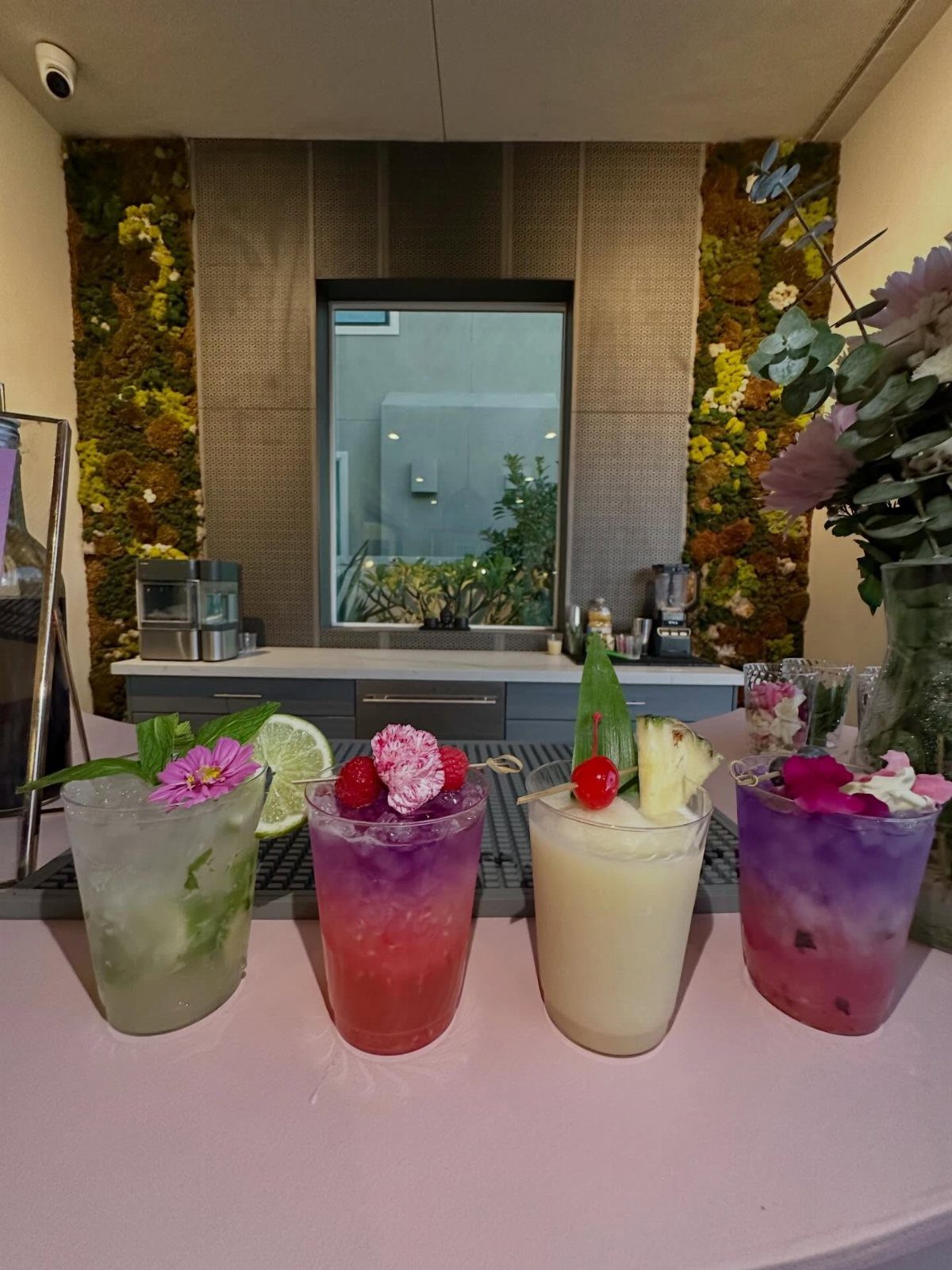 Four colorful garnished mocktails lined up on a pink counter in a modern room.