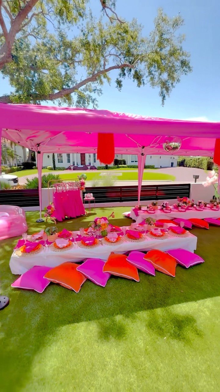 Pink outdoor party setup with long tables, floor cushions, and a pink canopy.