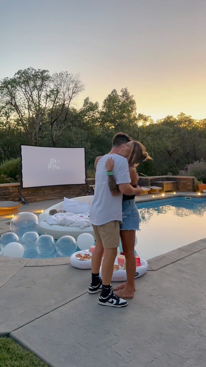 Couple hugging by a pool with an outdoor movie projector screen and lounge seating.