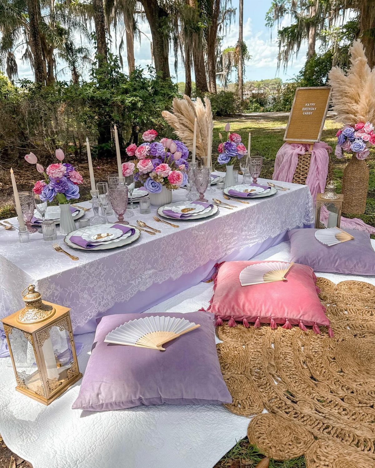 Low picnic table with lace cloth, purple flowers, and colorful cushions outdoors.