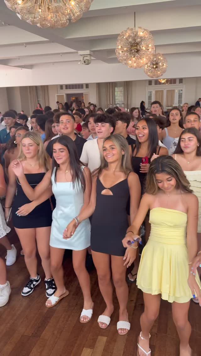 Teenagers in party dresses dancing and socializing under large spherical chandeliers in a hall.