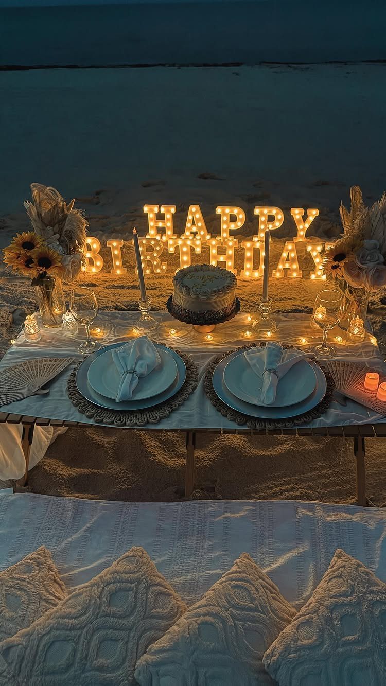 Beach birthday picnic setup with glowing letters, cake, and white pillows on sand.