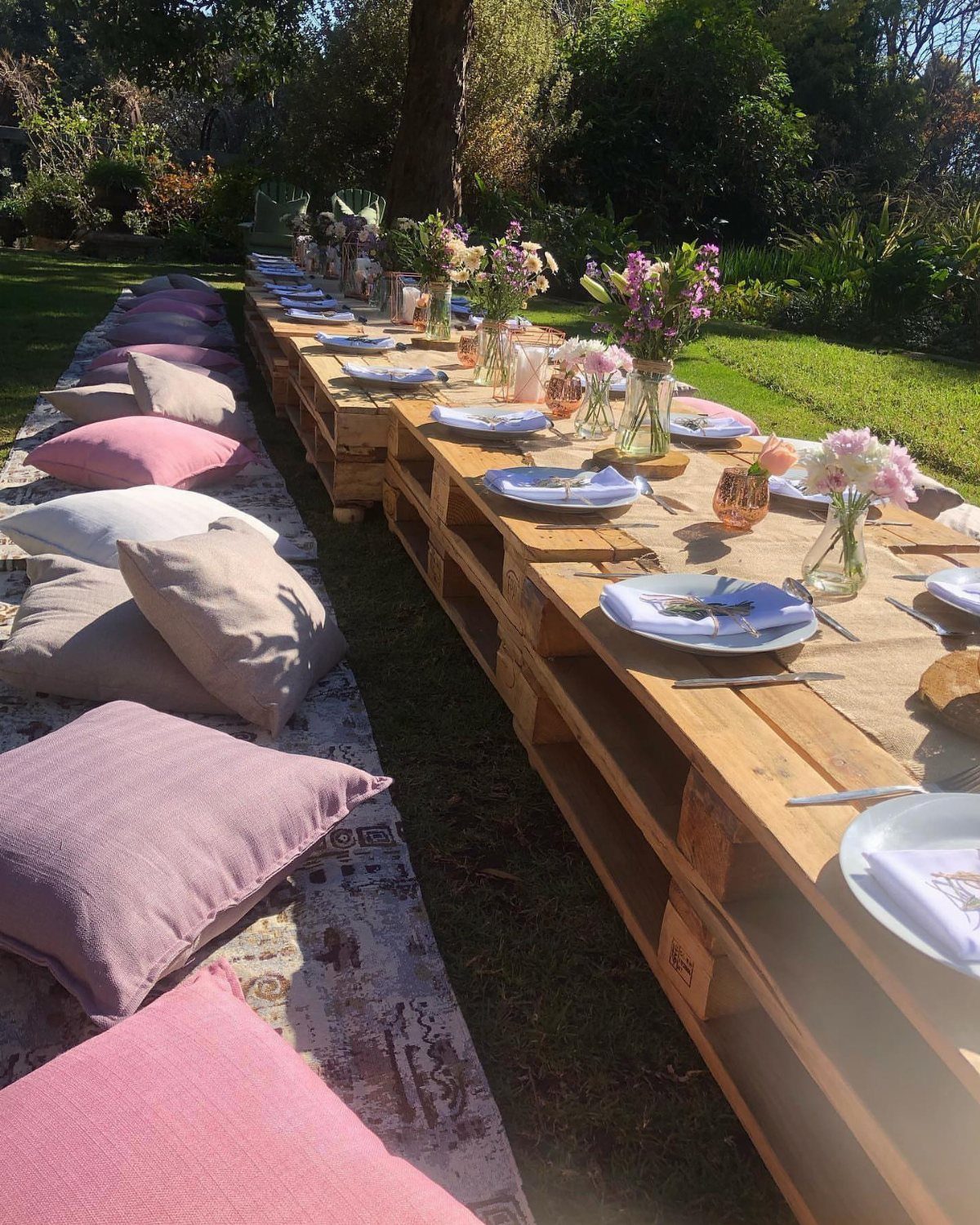 Wooden pallet picnic table with floral arrangements and pillows on a garden rug