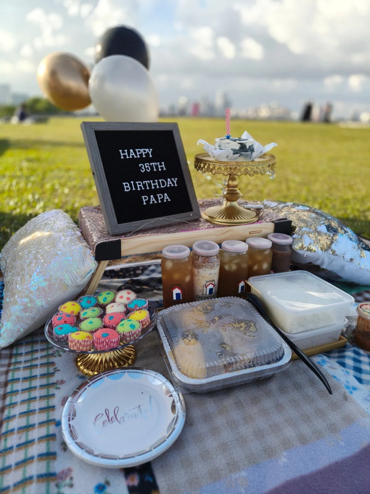 Birthday picnic setup with cupcakes, food containers, and a birthday sign on grass.