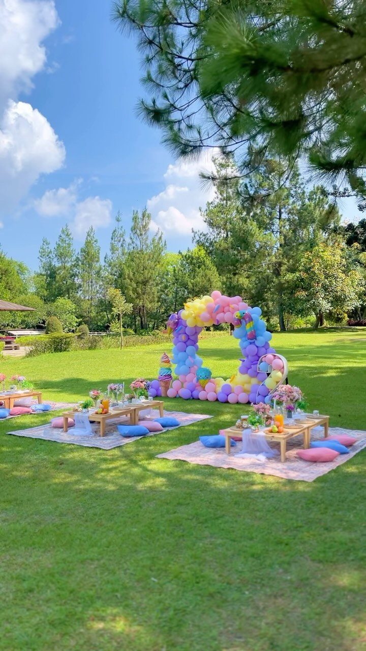 Outdoor picnic setup with low tables, colorful pillows, and a large rainbow balloon arch.
