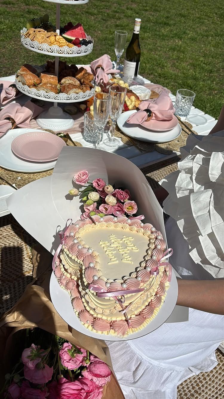 Heart-shaped pink cake held in front of a lavish outdoor picnic spread on grass