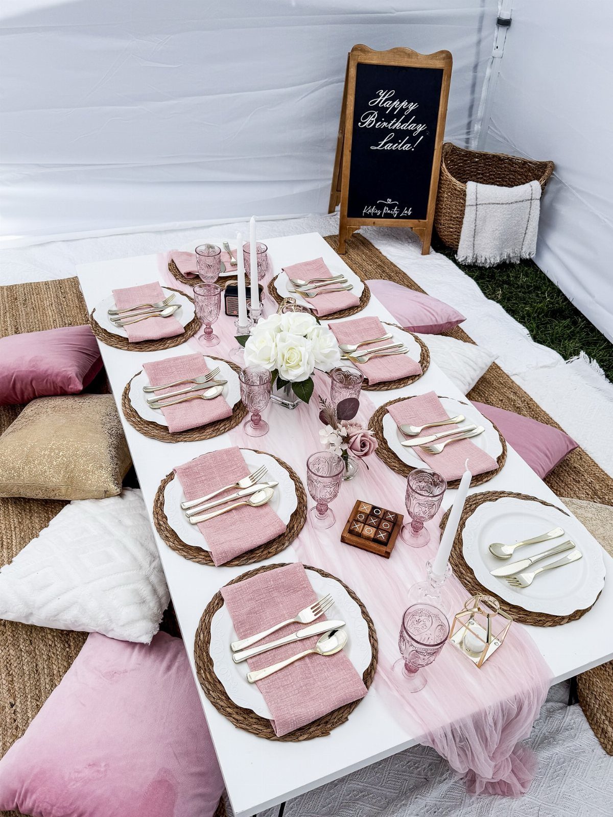 Low white picnic table set with pink glassware, plates, and floral centerpieces.