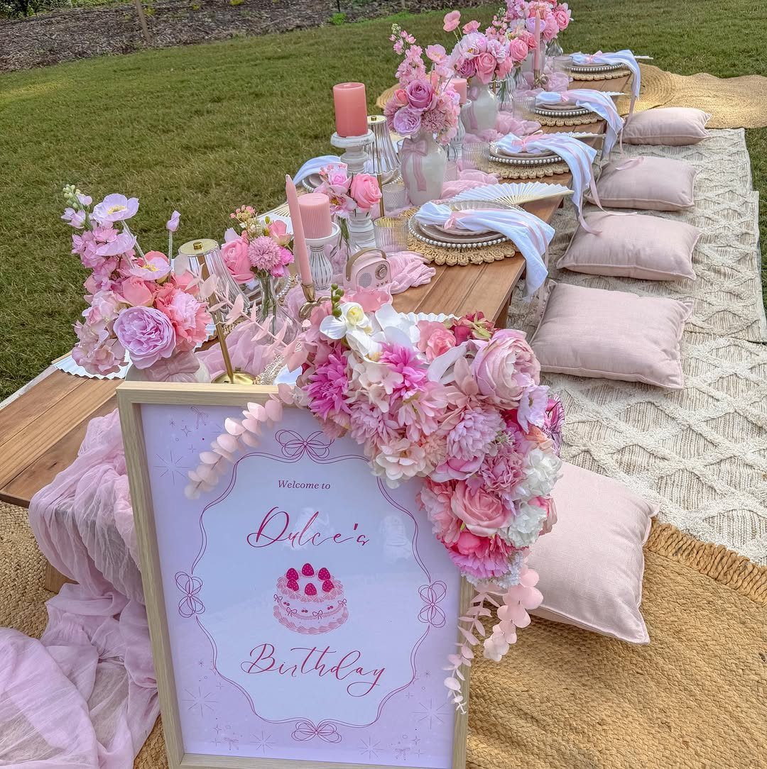 Low wooden picnic table with pink floral arrangements and pillows on a tapestry blanket.