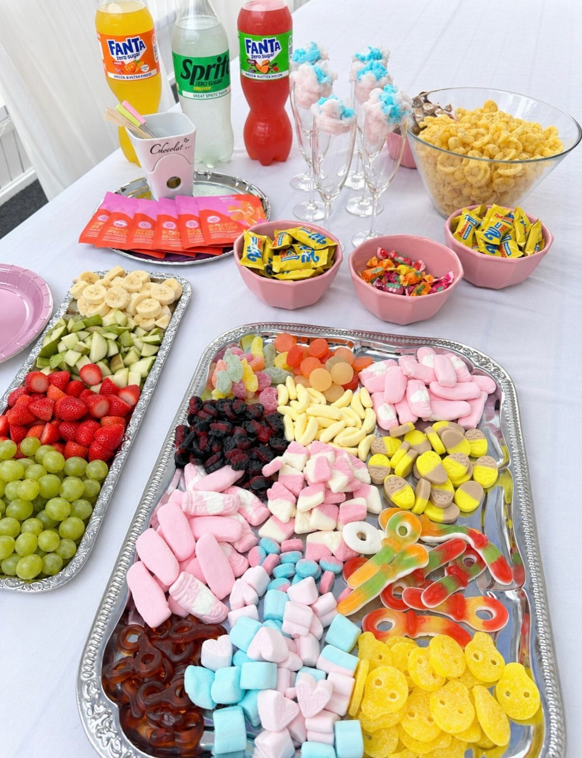 Assorted gummy candies, marshmallows, fresh fruit, and sodas arranged on a white table.