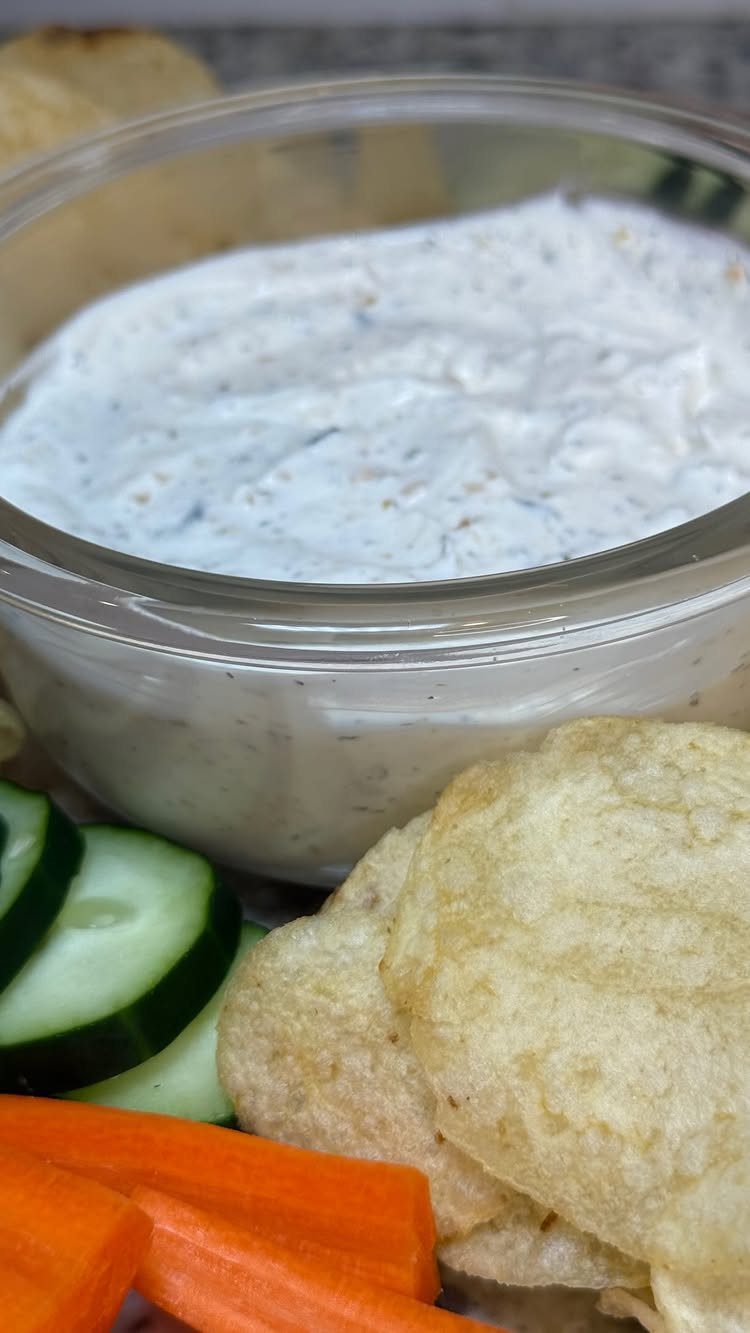 White creamy dip in a glass bowl with potato chips and fresh vegetables.